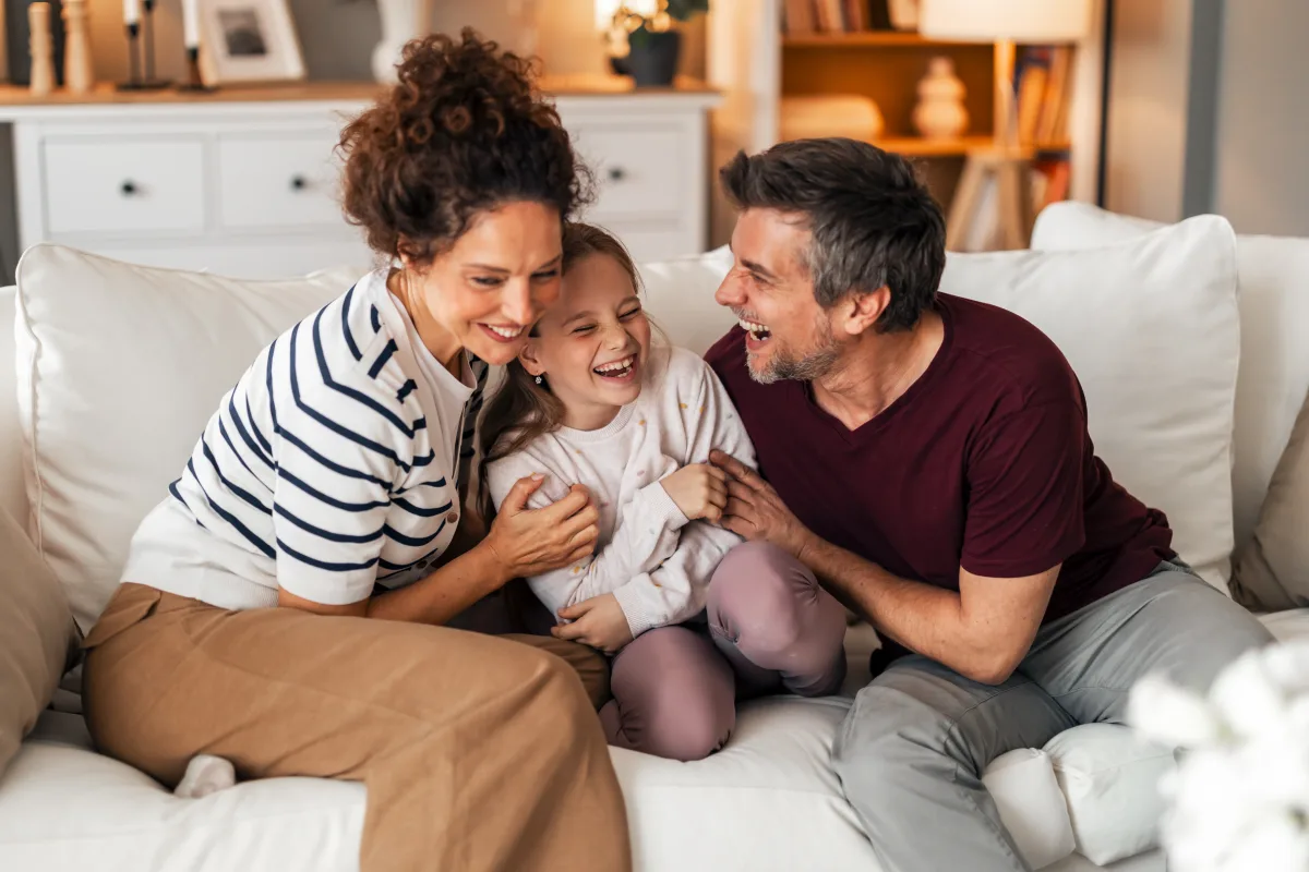 Smiling parents laughing and hugging their happy young daughter on a white couch in a cozy living room.