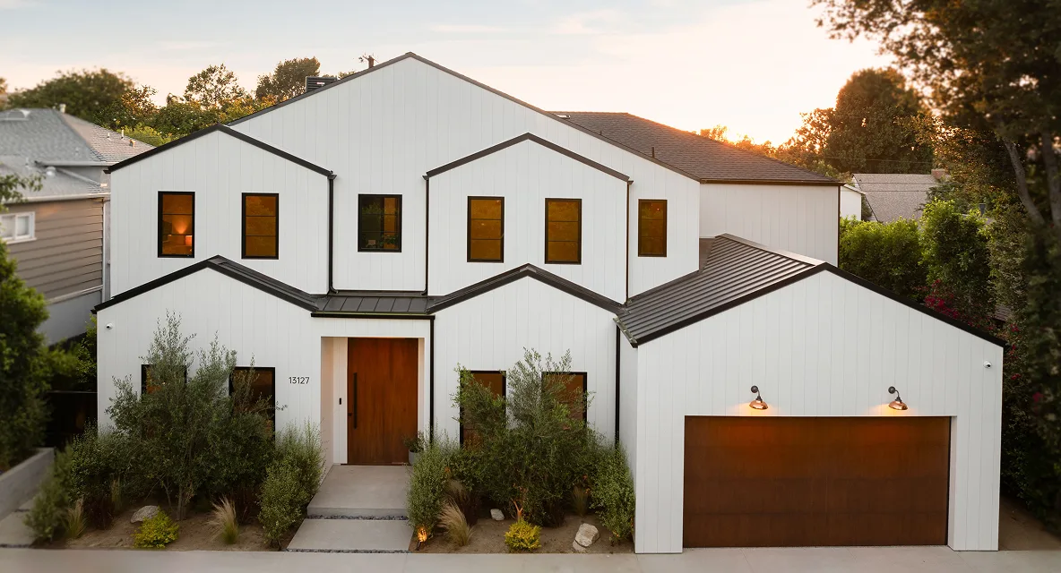 Two-story modern white house with black trim, wooden front door, large garage, and surrounding greenery at sunset.